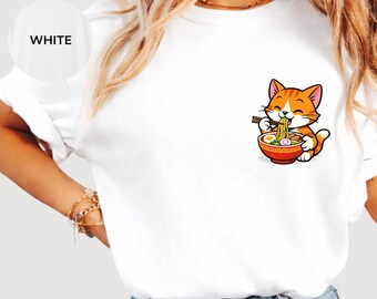 May include: A white t-shirt featuring a graphic print on the left chest area. The illustration depicts an orange tabby cat eating a bowl of ramen noodles with chopsticks. The bowl is red and contains noodles, broth, and egg slices.