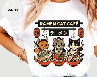 May include: A white t-shirt featuring a graphic of three cats eating ramen in bowls. The text reads RAMEN CAT CAFÉ with Japanese characters on hanging lanterns. The cats are orange, gray, and black and white, each wearing an apron and holding utensils.