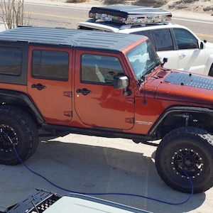 Peut inclure: Un Jeep Wrangler orange vif avec un toit rigide noir, des roues noires et un câble de remorquage bleu. Le Jeep est garé sur un chemin de terre avec un SUV blanc en arrière-plan.
