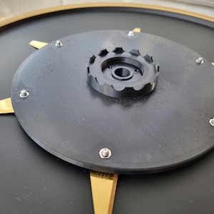 May include: A black and gold clock face with a central black disc secured by silver screws. A smaller, black, gear-shaped component sits atop the disc. Gold-colored, triangular accents radiate from the center, creating a modern, geometric design.