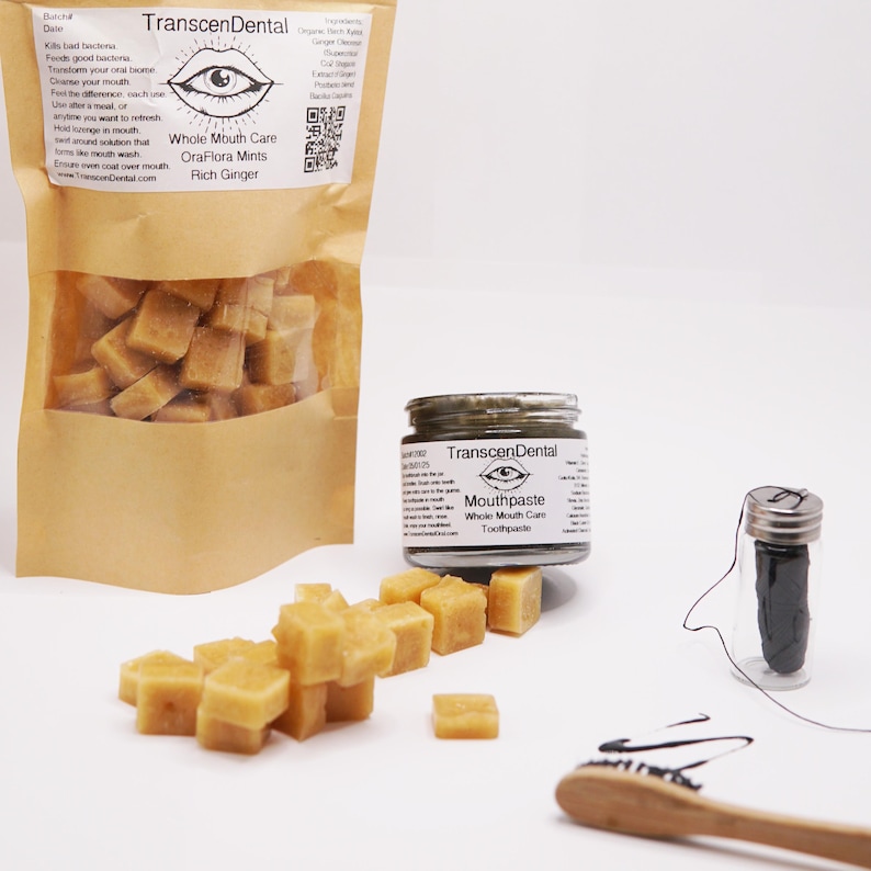 May include: A display of oral care products, including a bag of OraFlora Mints, a jar of toothpaste, a container of dental floss, and a wooden toothbrush. The mints are small, square, and light brown. The toothpaste jar has a silver lid and a label. The floss is black.