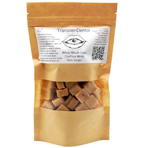 Op de afbeelding: Een bruine papieren zak met een doorzichtig venster, gevuld met vierkante, lichtbruine pepermuntjes. De zak heeft een wit etiket met de tekst "TranscenDental" en "OraFlora Mints Rich Ginger". Het etiket bevat ook een afbeelding van een oog en een mond.