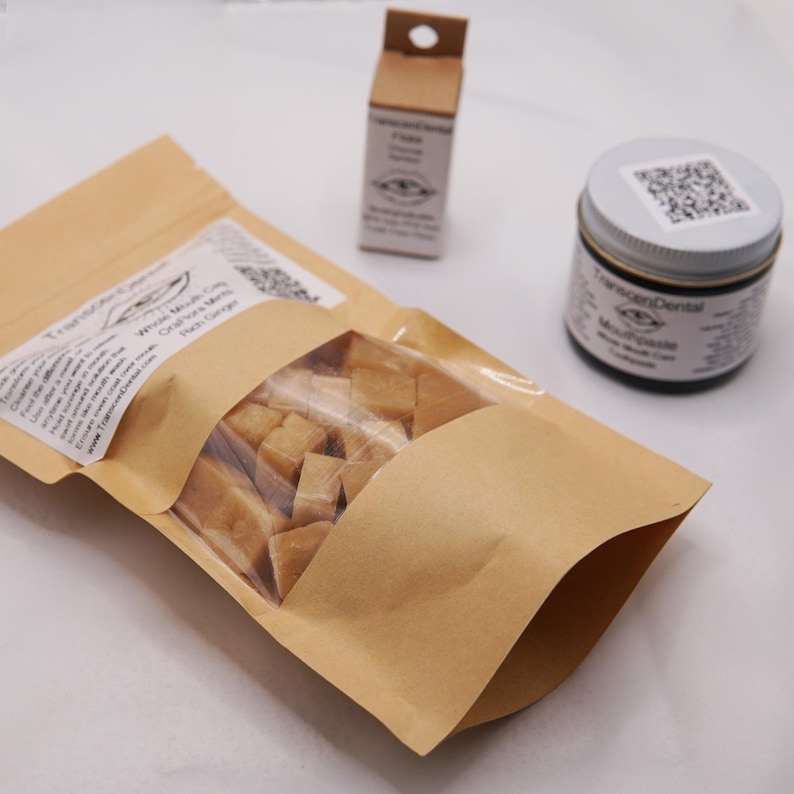 May include: A collection of dental care products, including a bag of brown, square-shaped items, a small box, and a jar with a silver lid. The bag has a clear window and a label with text. The jar and box also have labels. The products are arranged on a white surface.