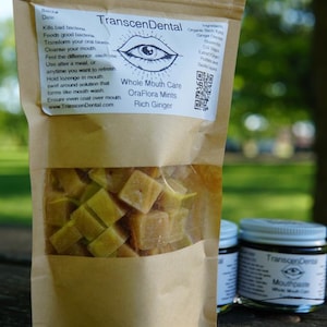 May include: A brown paper bag with a clear window displaying square, yellow-green mints. The bag is labeled "TranscenDental OraFlora Mints Rich Ginger." A jar of mouthpaste and a toothbrush are also visible.