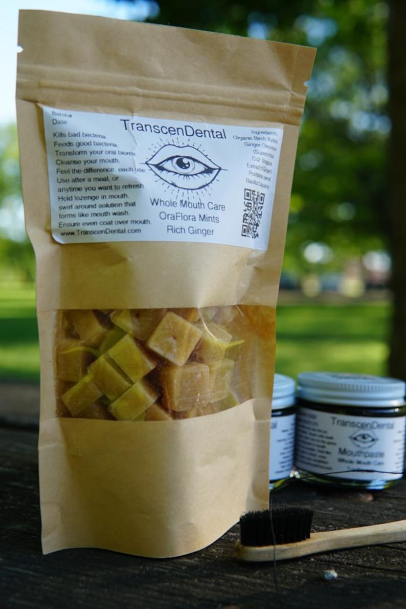 May include: A brown paper bag with a clear window displaying square, yellow-green mints. The bag is labeled "TranscenDental OraFlora Mints Rich Ginger." A jar of mouthpaste and a toothbrush are also visible.