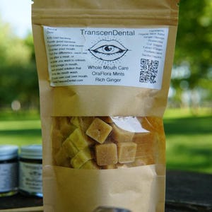 May include: A brown paper bag of OraFlora Mints, a whole mouth care product. The bag has a clear window showing the square-shaped mints. The label reads "TranscenDental Whole Mouth Care OraFlora Mints Rich Ginger."