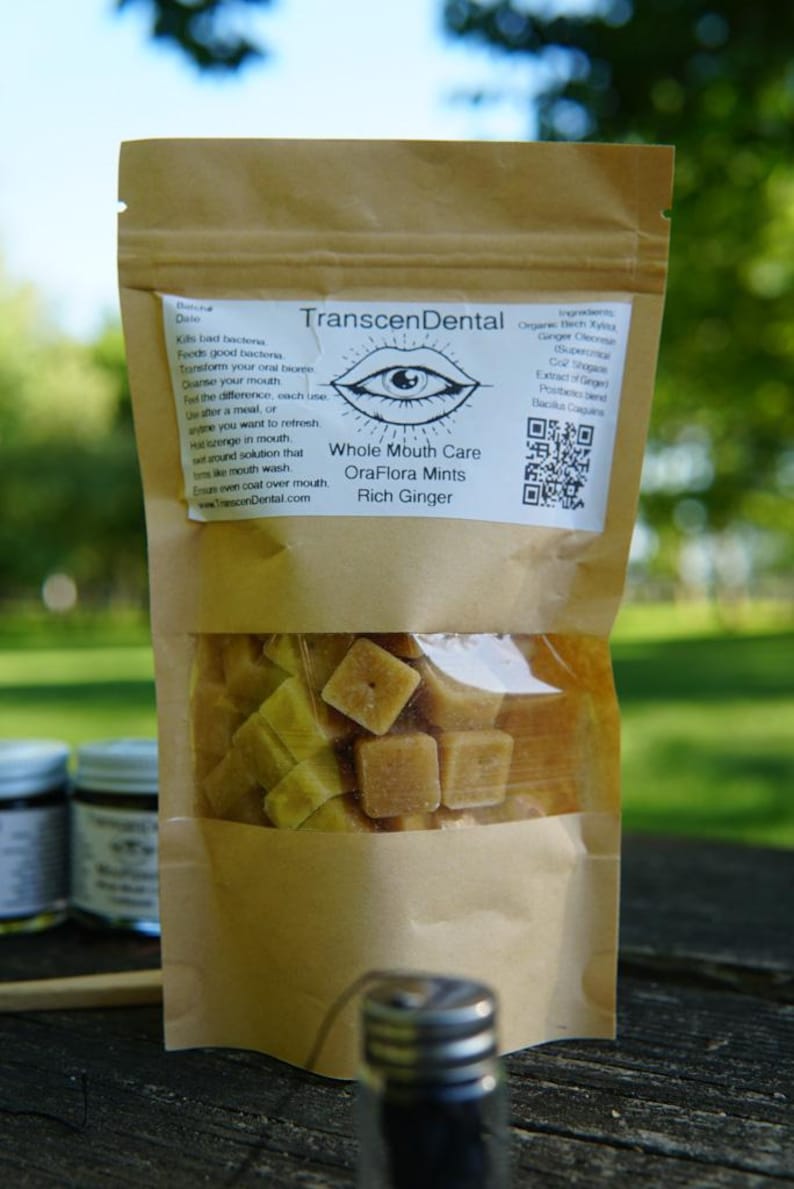 May include: A brown paper bag of OraFlora Mints, a whole mouth care product. The bag has a clear window showing the square-shaped mints. The label reads "TranscenDental Whole Mouth Care OraFlora Mints Rich Ginger."