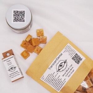 May include: An overhead shot of dental care products. A small tin with a QR code, a box of floss, and a brown paper bag with a label that reads "Transcendental" and "OraFlora Mints". Scattered around are small, square, tan-colored mints.