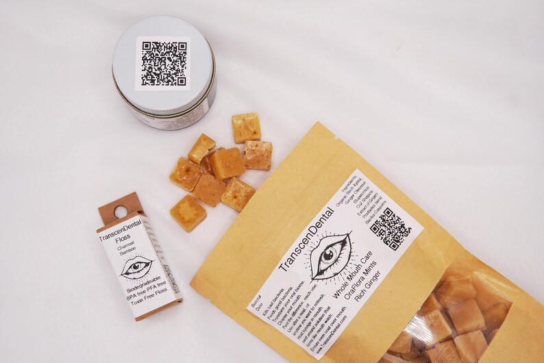 May include: An overhead shot of dental care products. A small tin with a QR code, a box of floss, and a brown paper bag with a label that reads "Transcendental" and "OraFlora Mints". Scattered around are small, square, tan-colored mints.