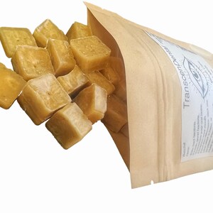 May include: A close-up shot shows a collection of light yellow, cube-shaped items spilling from a brown paper bag. The bag has a white label with the text "Transcendental Dental" and an eye graphic. The cubes appear to be a dental product.