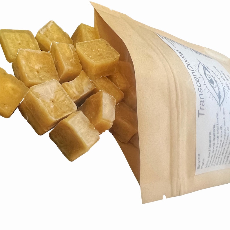 May include: A close-up shot shows a collection of light yellow, cube-shaped items spilling from a brown paper bag. The bag has a white label with the text "Transcendental Dental" and an eye graphic. The cubes appear to be a dental product.