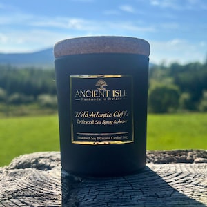 Peut inclure: Un pot de bougie noir avec un couvercle en liège. L'étiquette indique "ANCIENT ISLE Handmade in Ireland" et "Wild Atlantic Cliffs" avec des notes de parfum. La bougie est sur une surface en bois avec un fond vert.