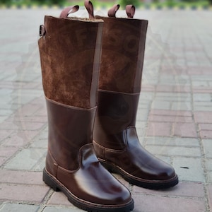 Pode incluir: Um par de botas altas de couro castanho com um acabamento brilhante. As botas têm uma parte superior em camurça e uma fivela. As solas são pretas e as botas estão sobre uma superfície de tijolo. O texto "SwiftSoleLeather" está na parte inferior.