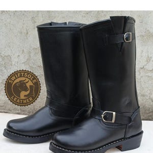 Könnte beinhalten: Schwarze Lederstiefel mit rundem Zehenbereich und mittelhohem Schaft. Die Stiefel haben einen Schnallenriemen um den Knöchel und einen kleinen Absatz. Das Swiftsole Leather-Logo ist sichtbar.