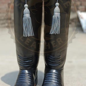 Könnte beinhalten: Ein Paar hohe, schwarze Lederstiefel mit silbernen Verzierungen am oberen Rand und dekorativen silbernen Quasten. Die Stiefel haben ein klassisches Design mit einem geriffelten Muster am Unterschenkel und einer runden Spitze. Der Markenname "SwiftSoleLeather" ist sichtbar.