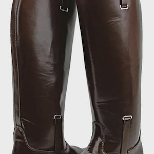 May include: A pair of tall, brown leather riding boots. The boots have a glossy finish, a pull tab at the top, and a strap with a buckle near the ankle. The boots are designed for equestrian activities.