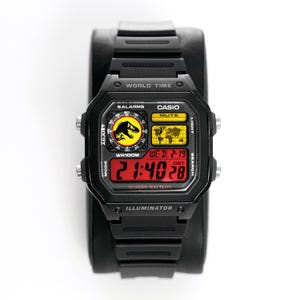 Puede incluir: Reloj digital Casio negro con esfera rectangular. El reloj muestra la hora en dígitos rojos, con la fecha y un gráfico de mapa del mundo. El reloj tiene el texto "WORLD TIME", "CASIO" e "ILLUMINATOR". La correa del reloj es negra.