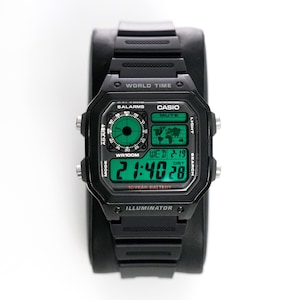 Puede incluir: Reloj digital Casio negro con esfera rectangular y correa negra. La esfera muestra la hora, la fecha y un mapa del mundo. El reloj tiene el texto "WORLD TIME", "CASIO" e "ILLUMINATOR".