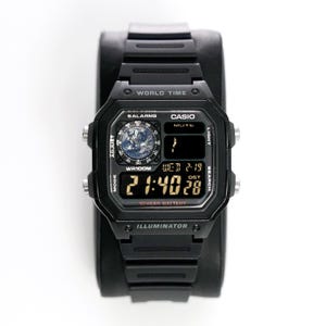 May include: Black digital watch with a square face and black band. The watch face displays the time, date, and world time features. The watch has the text "CASIO" and "ILLUMINATOR" printed on the face.