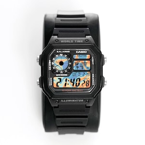 Puede incluir: Reloj digital Casio negro con esfera cuadrada. El reloj muestra la hora, la fecha y las funciones de hora mundial. El reloj tiene una correa negra y está colocado sobre un soporte negro. La esfera del reloj tiene el texto "CASIO" e "ILLUMINATOR".