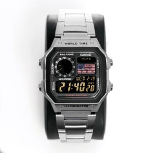 Könnte beinhalten: Eine silberfarbene Casio Digitaluhr mit schwarzem Zifferblatt und Edelstahlarmband. Die Uhr zeigt Uhrzeit, Datum und eine amerikanische Flagge an. Funktionen: Weltzeit, Alarme und 10-Jahres-Batterie.