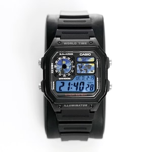 May include: Black Casio digital watch with a square face and black band. The watch displays the time, date, and world time features. The face includes the text "CASIO", "WORLD TIME", and "ILLUMINATOR". The watch is displayed on a black stand.