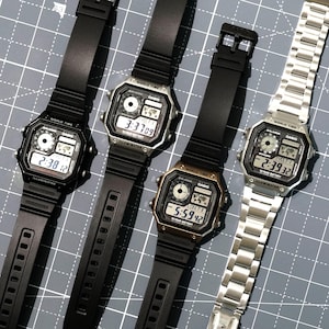 May include: Four digital watches with black and silver cases and bands. One watch has a silver band. The watch faces display the time and world time features. The watches are on a gray surface.