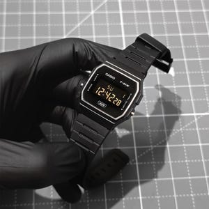 K&ouml;nnte beinhalten: Eine schwarze Casio F-91W Digitaluhr mit rechteckigem Zifferblatt und schwarzem Metallarmband. Die Uhr zeigt die Uhrzeit 12:42:28 an. Die Uhr wird von einer behandschuhten Hand vor einem grauen Gitterhintergrund gehalten.