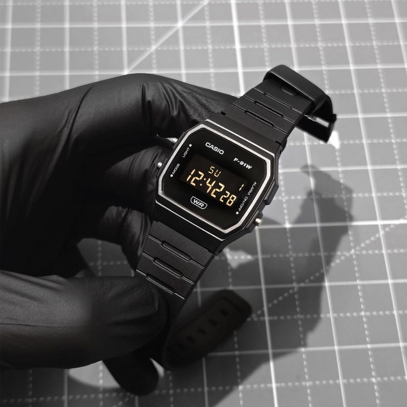 K&ouml;nnte beinhalten: Eine schwarze Casio F-91W Digitaluhr mit rechteckigem Zifferblatt und schwarzem Metallarmband. Die Uhr zeigt die Uhrzeit 12:42:28 an. Die Uhr wird von einer behandschuhten Hand vor einem grauen Gitterhintergrund gehalten.