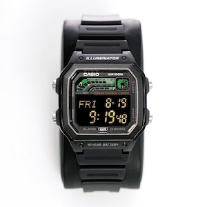 Peut inclure: Montre numérique Casio noire avec un cadran rectangulaire. La montre affiche la date et l'heure, avec le texte "ILLUMINATOR" au-dessus de l'écran. La montre a un bracelet noir et est présentée sur un support noir.