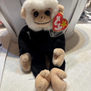 Puede incluir: Un mono de peluche Beanie Baby negro y blanco con la cara blanca y el cuerpo negro. El mono está sentado sobre una superficie blanca y tiene una etiqueta Ty roja y blanca unida a su oreja. La etiqueta dice "Beanie Babies" y tiene una estrella.