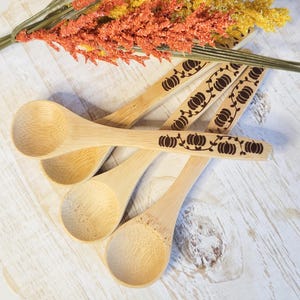 May include: A collection of bamboo spoons with pumpkin designs. The spoons are light brown with a carved pumpkin and vine pattern on the handles. The spoons are arranged on a light-colored wooden surface, with orange and yellow decorative foliage.