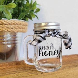 May include: A small, clear glass jar with a handle and a silver lid. The jar is decorated with a black and white checkered ribbon tied around the lid. The words "farm honey" are printed on the front of the jar.