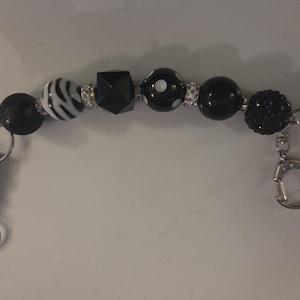 May include: A beaded keychain featuring a variety of black and white beads. The beads include a zebra-striped bead, a faceted bead, and several solid black beads. Silver-colored metal clasps are attached at each end.