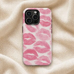 May include: A phone case with a pattern of pink lipstick kiss marks on a white background. The case has a black border and a camera cutout. The background is a beige fabric.