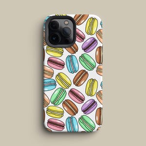 May include: A white phone case featuring a repeating pattern of colorful macarons. The macarons are in various colors, including yellow, pink, blue, green, brown, and purple, with a detailed, cartoon-like design. The case has a camera cutout.