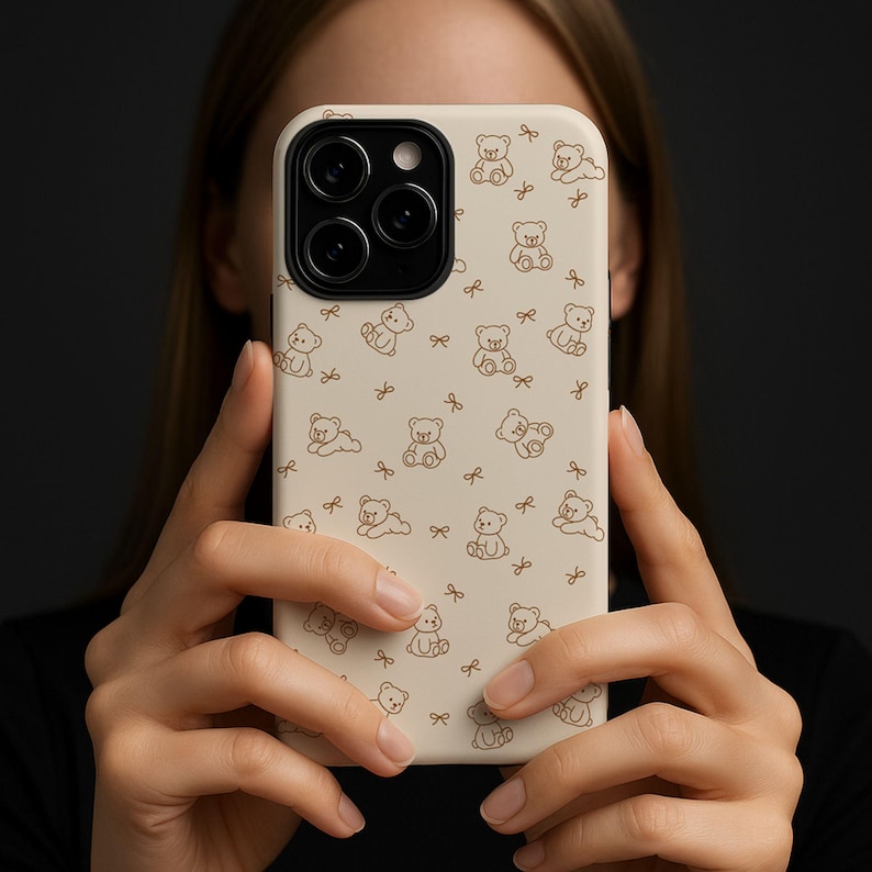 May include: A beige phone case featuring a repeating pattern of brown teddy bears and bows. The bears are in various poses, and the bows are scattered throughout the design. The case is held in front of a person.