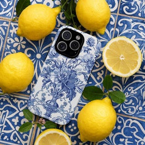 Könnte beinhalten: Ein weiß-blaues Handycase mit detailliertem Blumenmuster, umgeben von frischen gelben Zitronen und grünen Blättern. Das Case hat ein klassisches blau-weißes Muster, das an Porzellan erinnert. Die Zitronen sind ganz und geschnitten, was einen lebendigen Kontrast erzeugt.