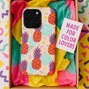 May include: A phone case with a pineapple design in a gift box. The case has a cream background with orange pineapples, teal circles, and pink accents. The box contains colourful tissue paper and a card that reads "MADE FOR COLOR LOVERS."