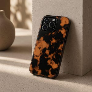 Puede incluir: Funda negra para smartphone con un diseño de carey naranja y negro. La funda tiene un acabado brillante y un módulo de cámara negro con tres lentes. La funda está apoyada contra un bloque de color neutro.