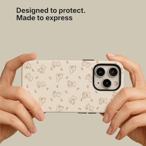 May include: A beige phone case with a repeating pattern of brown teddy bears and bows. The case has a black camera lens border and the text "Designed to protect. Made to express."