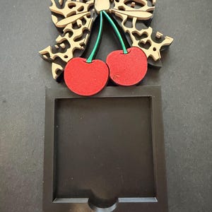 May include: A black square frame with a cutout at the bottom center, topped with a decorative bow featuring a tan, intricate, laser-cut pattern. Two bright red cherries with green stems are attached to the center of the bow, creating a contrast.