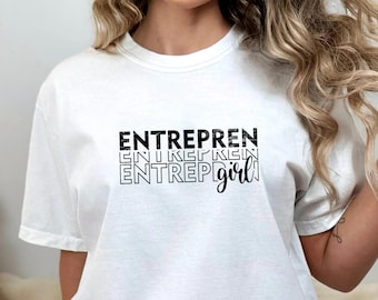 May include: White t-shirt with the word "ENTREPREN" stacked three times in a black, textured font, with "girl" in a script font. The shirt is a casual style.