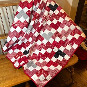 May include: A patchwork quilt featuring a repeating square pattern in shades of red, white, and black. The fabric squares display various prints including gingham, stars, and floral motifs, all joined by intricate white thread quilting patterns.