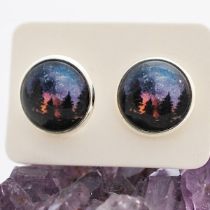 May include: A pair of silver stud earrings with a round, black background featuring a watercolor painting of a forest silhouette against a starry night sky.