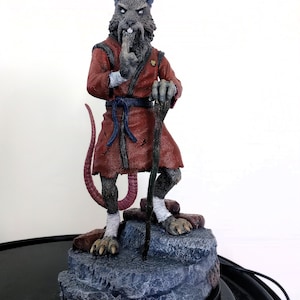 May include: A detailed figurine of a rat-like character in a red robe, holding a staff. The figure stands on a rocky base, with a long tail and a finger to its lips. The figure is approximately 7 inches tall.