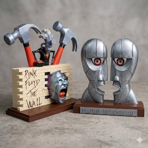 Puede incluir: Dos esculturas temáticas de Pink Floyd. Una presenta un muro de ladrillos con el texto "Pink Floyd The Wall", una cara gritando y martillos. La otra representa dos cabezas plateadas con ojos rojos, sobre una base que dice "Pink Floyd - The Division Bell".