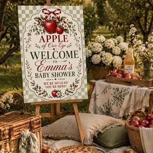 May include: A rectangular welcome sign for a baby shower featuring a light green checkered background, red apples, white flowers, and a red ribbon. The text reads: APPLE of Our Eye WELCOME TO Emma's BABY SHOWER WE'RE SO GLAD YOU'RE HERE!