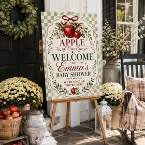 May include: A rectangular welcome sign for a baby shower featuring a light green and white checkered border. The sign displays the text APPLE of Our Eye WELCOME TO Emma's BABY SHOWER WE'RE SO GLAD YOU'RE HERE! with red apple and floral illustrations.