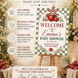 May include: A rectangular sign on a wooden easel featuring a green and white checkered border, red apples, white blossoms, and a red ribbon. The text reads: Welcome to Emma's Baby Shower, We're so glad you're here! The sign includes instructional text.